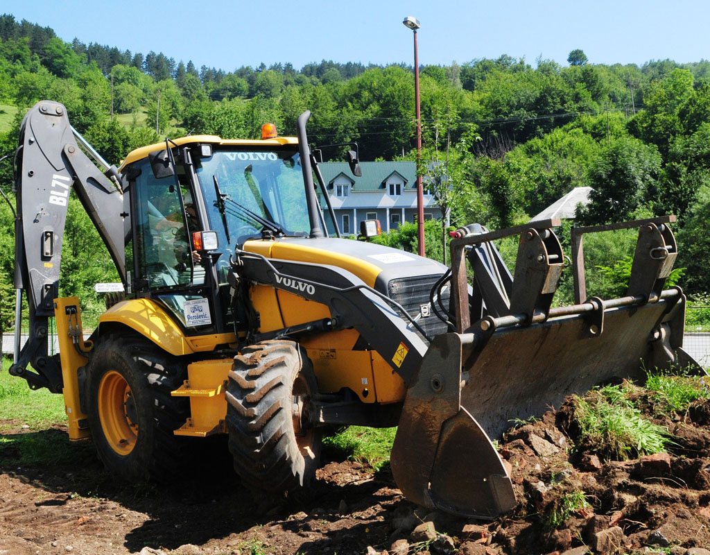 Backhoe moving dirt and stones on a lawn, with a house and trees in the background