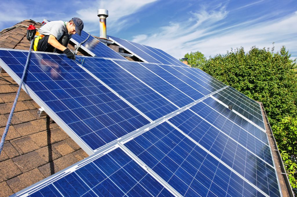  Man installs solar panels on a roof with a blue sky and trees in the background.