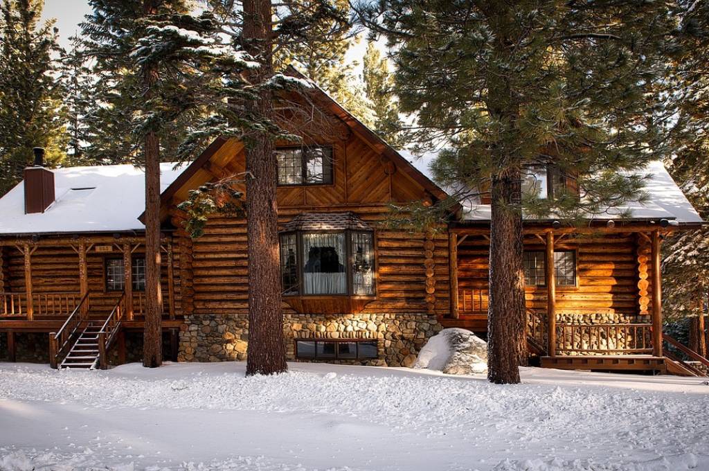 Large pine trees stand tall in front of a large, brown log cabin with a snow-covered roof in a snowy forest.