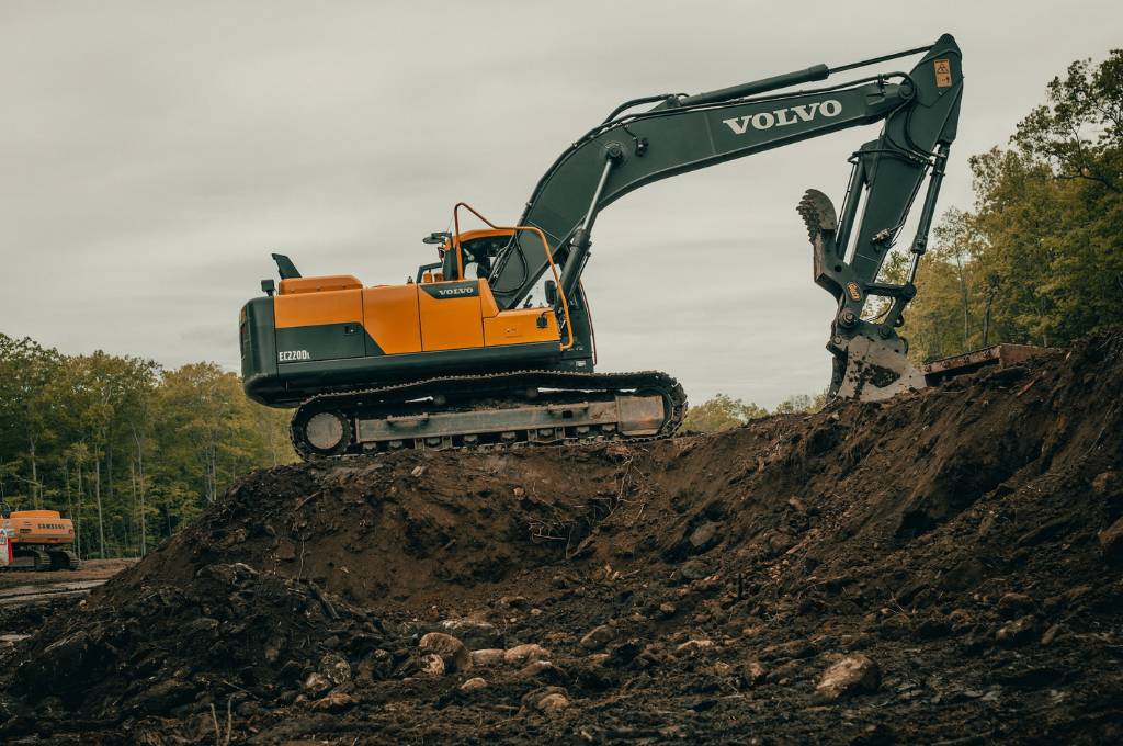 A yellow and black Volvo backhoe excavator uses its arm and shovel to dig up soil in a forested area.