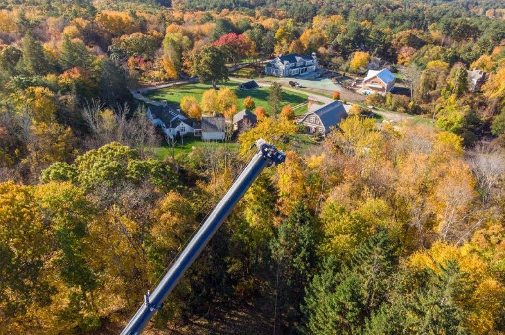 An aerial view of a crane arm extending over a wooded, fall-colored residential neighborhood during a tree removal.