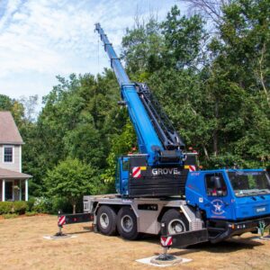 The blue American Climbers tree removal crane sits parked in the front yard of a gray suburban two-story home with its boom extended and four outriggers set laterally to support the weight of the removal.